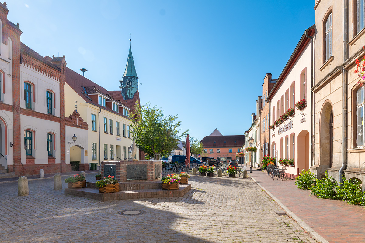 Marktplatz in Krakow am See mit historischen Gebäuden, Brunnen und Außengastronomie bei sonnigem Wetter.