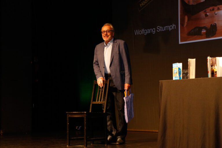 Schauspieler Wolfgang Stumph steht auf einer Bühne bei einer AWG-Buchlesung in Güstrow, neben ihm ein Tisch mit Büchern und Beleuchtung.